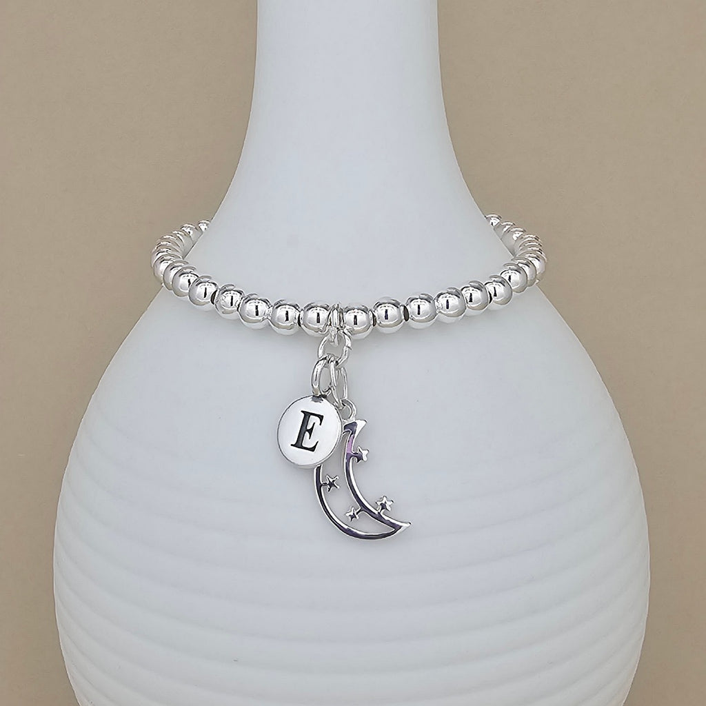 Silver bracelet with a moon charm and letter 'E' on a white vase.