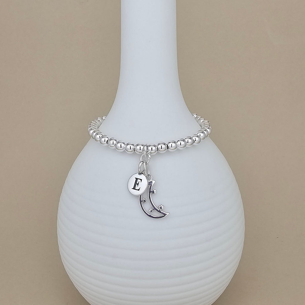 A silver beaded bracelet with a crescent moon and initial 'E' charm, displayed on a white ribbed ceramic vase against a beige background.