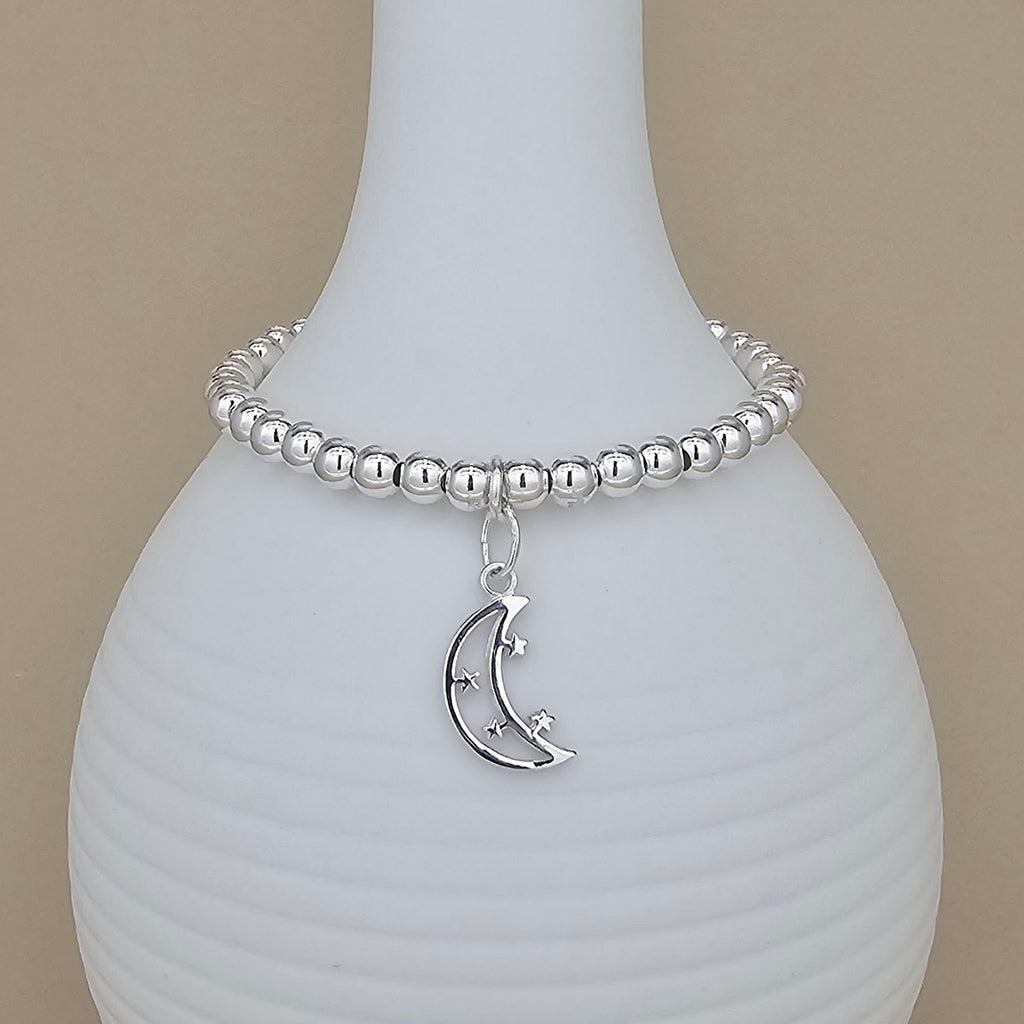A silver beaded bracelet with a crescent moon and star charm, displayed on a white ribbed ceramic vase against a beige background.
