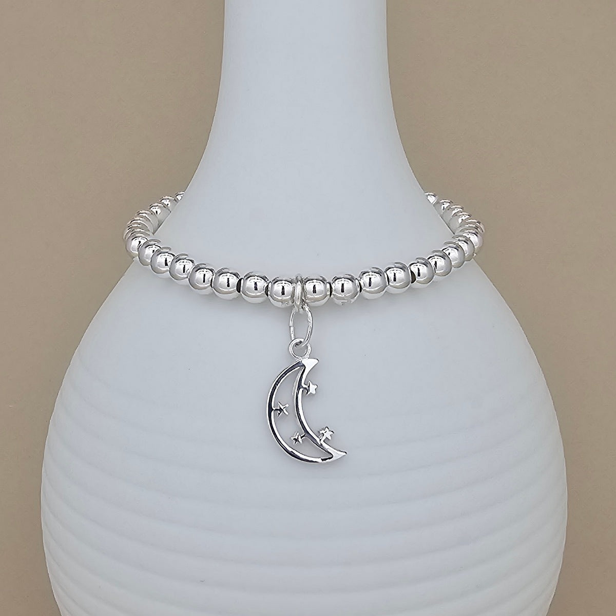 A silver beaded bracelet with a crescent moon and star charm, displayed on a white ribbed ceramic vase against a beige background.