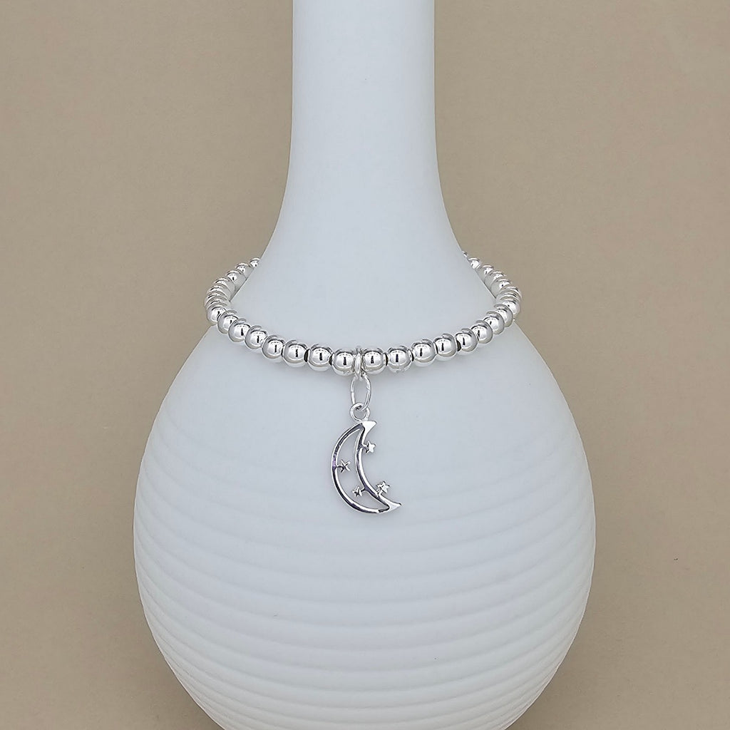 A silver beaded bracelet with a crescent moon and star charm, displayed on a ribbed white ceramic vase against a solid beige background.