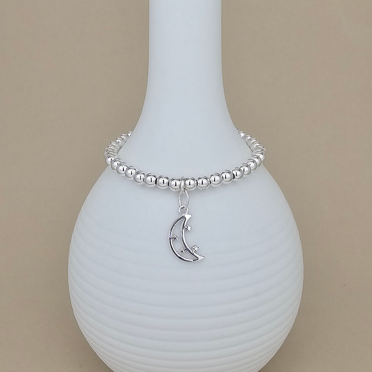 A silver beaded bracelet with a crescent moon and star charm, displayed on a ribbed white ceramic vase against a solid beige background.