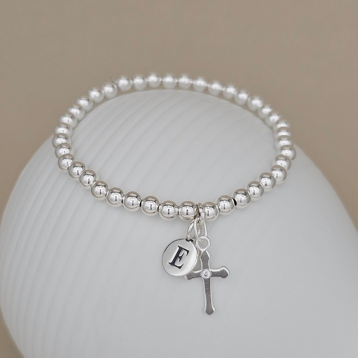 Silver beaded bracelet with a cross charm and letter 'D' charm on a white surface.