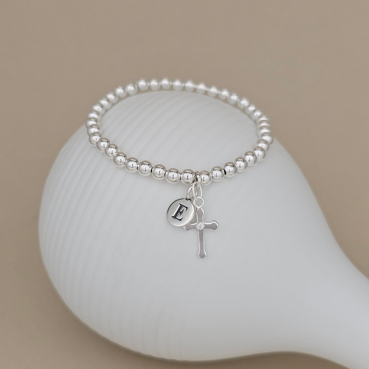 A silver beaded bracelet with a small initial 'E' charm and a cross charm, displayed on a white ceramic dish against a warm beige background.