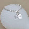 A silver beaded bracelet with a cross charm and a heart-shaped tag engraved with the name 'Poppy', displayed on a white ribbed shell against a beige background.