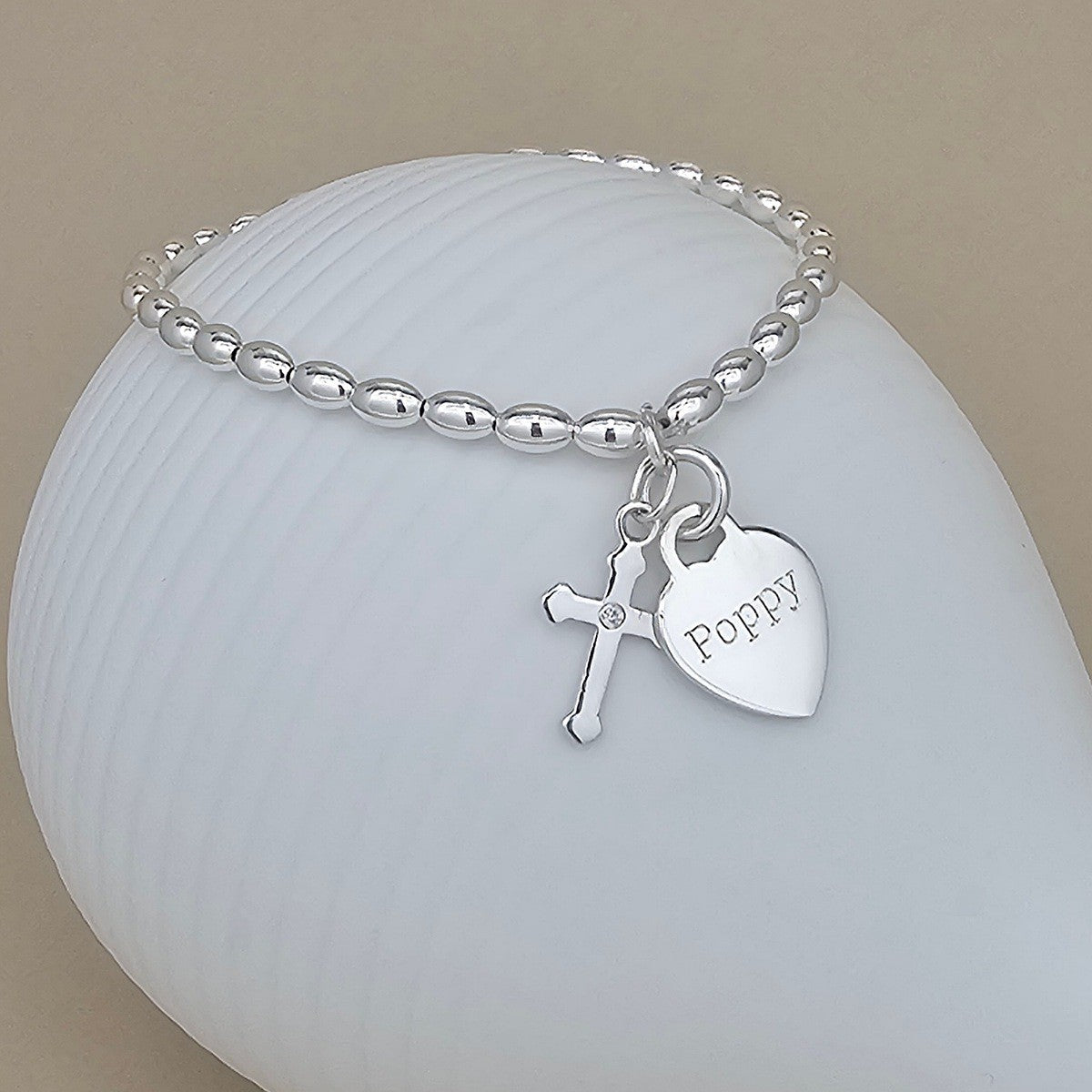 A silver beaded bracelet with a cross charm and a heart-shaped tag engraved with the name 'Poppy', displayed on a white ribbed shell against a beige background.
