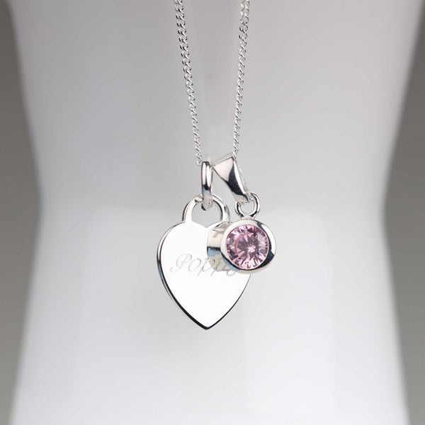 A silver heart-shaped pendant necklace with with engraved name and a small round pink gemstone charm, displayed on a white mannequin bust.