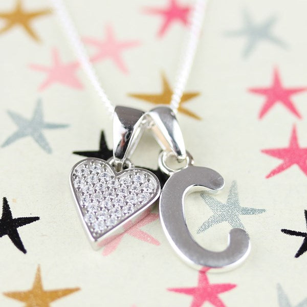A silver initial 'C' pendant necklace with a matching silver heart charm paved with small clear stones, hanging from a white cord against a light background with colorful star patterns.