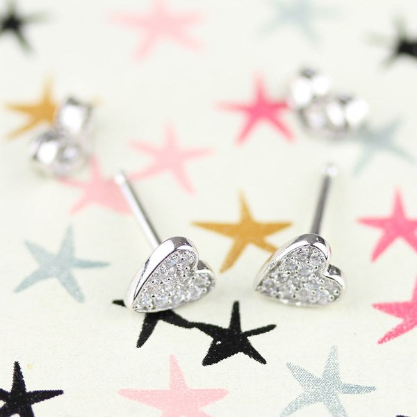 Silver heart-shaped stud earrings covered in clear rhinestones, displayed on a light cream background with scattered star stickers in pink, black, yellow, and blue.