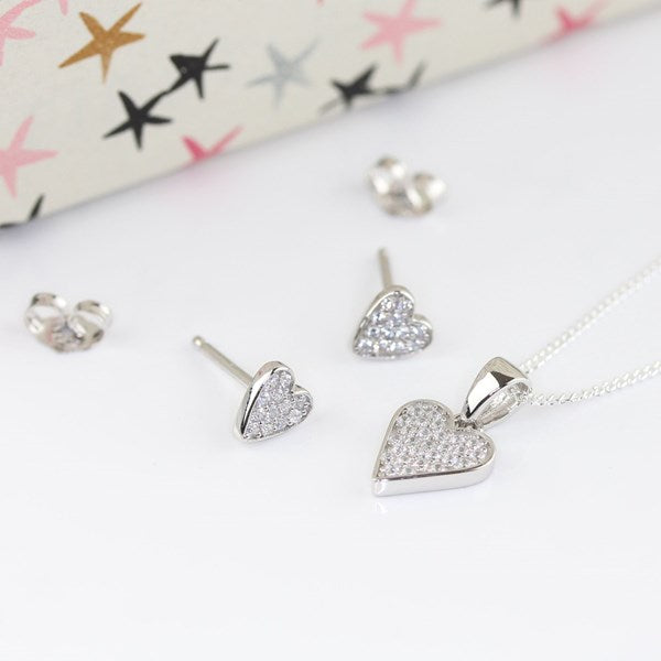 Silver heart jewelry set featuring a necklace with a pavé heart pendant, matching stud earrings, and heart-shaped backings, displayed on a white surface with a patterned gift box in the background.