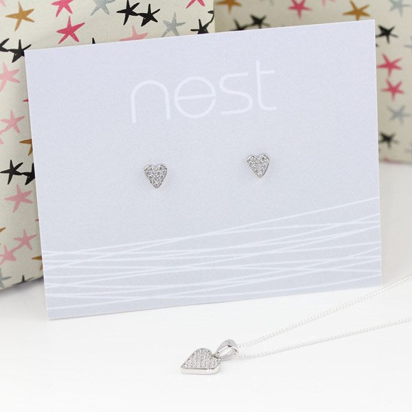 Silver heart stud earrings and matching heart pendant necklace on a white display card branded nest, with a white surface and patterned gift boxes in the background.