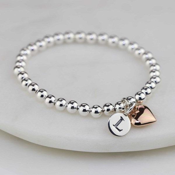 A silver beaded stretch bracelet with a polished silver disc charm engraved with the letter 'I' and a rose gold heart charm, displayed on a white marble surface.