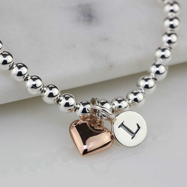 A silver beaded bracelet with a rose gold heart charm and a silver disc charm engraved with the letter 'I', resting on a white marble block.