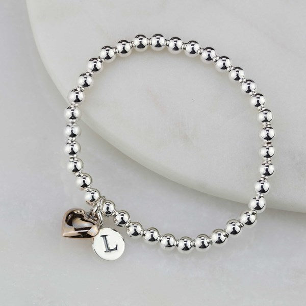 A silver beaded stretch bracelet with a dangling heart charm and a round initial 'L' charm, displayed on a white marble surface.