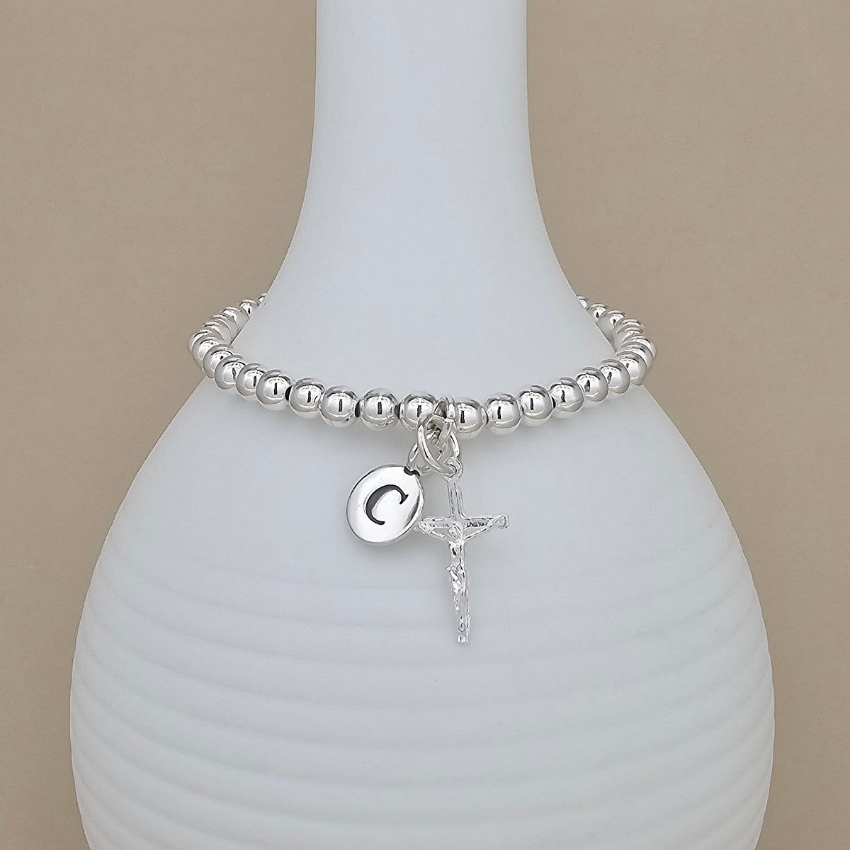 A silver beaded bracelet with a dangling initial 'C' charm and a small crucifix, displayed on a white ribbed vase against a beige background.