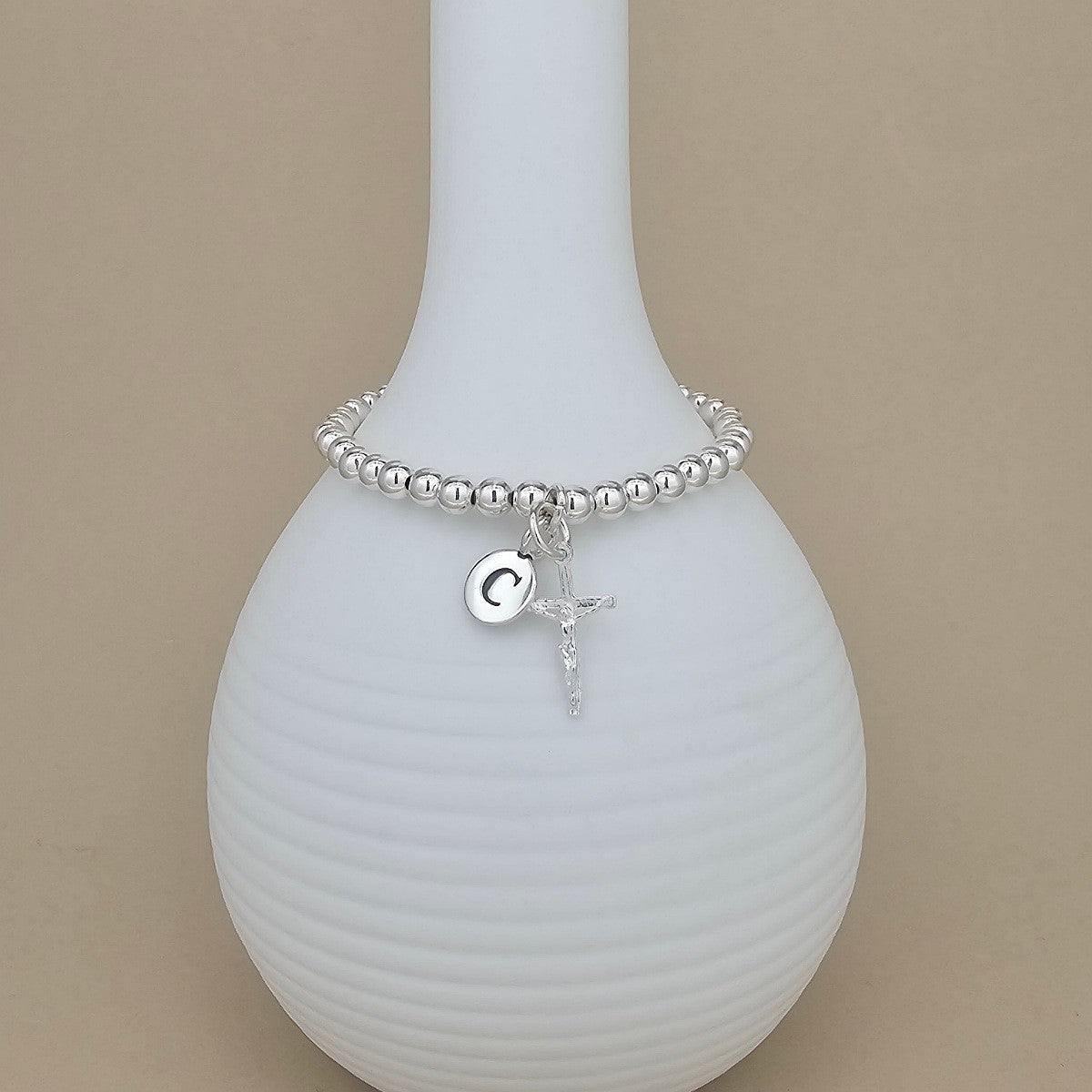A silver beaded bracelet with a circular initial 'G' charm and a small cross pendant, displayed on a white ribbed vase against a beige background.