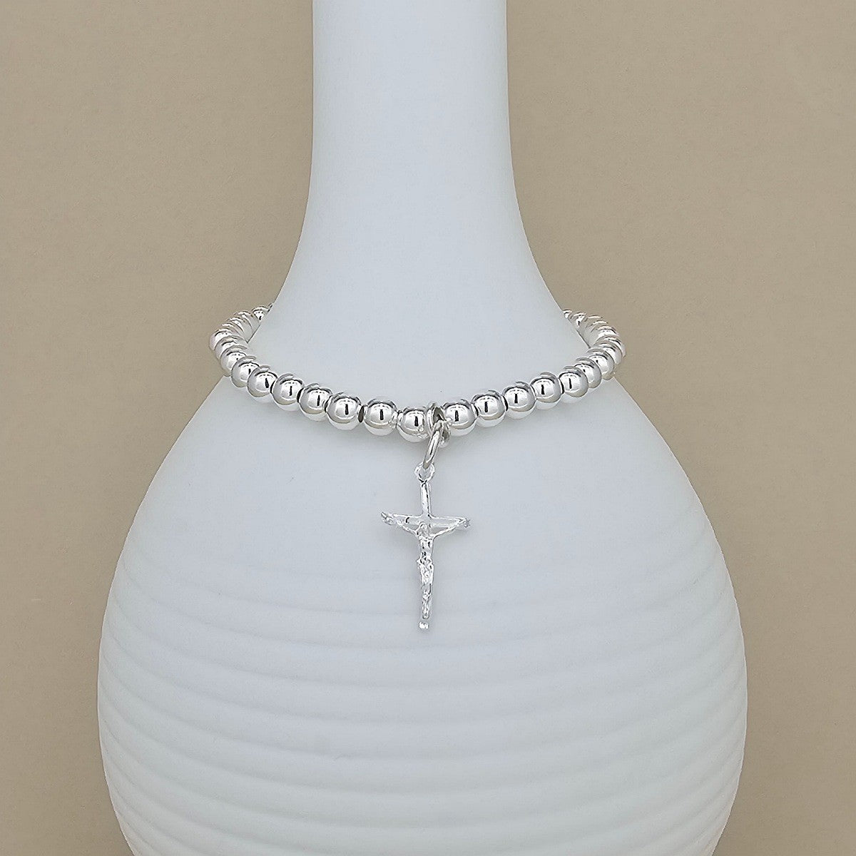 A silver beaded bracelet with a dangling crucifix charm, displayed on a white ribbed ceramic vase against a beige background.
