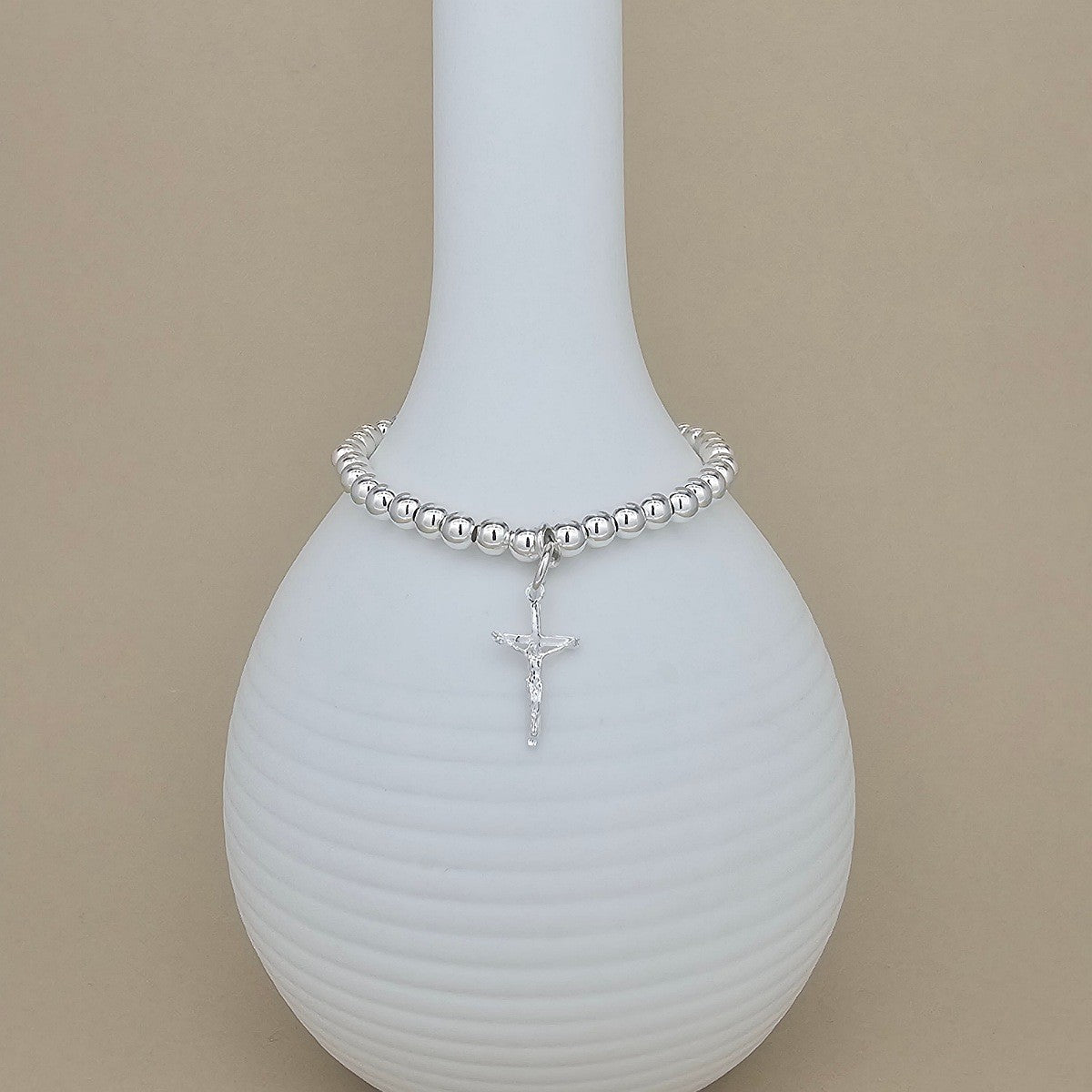 A silver beaded bracelet with a small crucifix pendant, displayed on a white ribbed vase against a beige background.