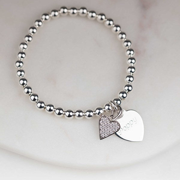 A silver beaded bracelet with two heart-shaped charms, one with cz stones and one engraved with the name 'poppy', displayed on a light marble surface.