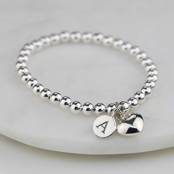 Silver beaded bracelet with a heart charm and round tag on a light gray background