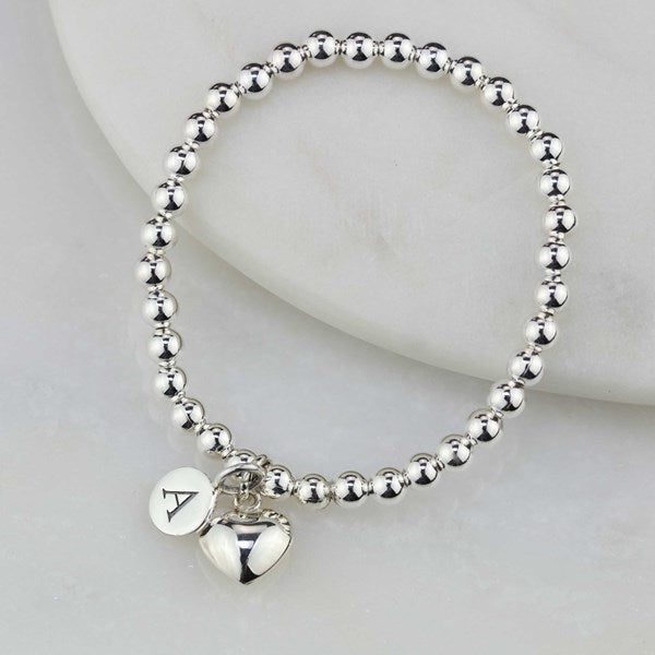 A silver beaded bracelet with heart charm and engraved disc with the letter 'A', displayed on a white marble surface.