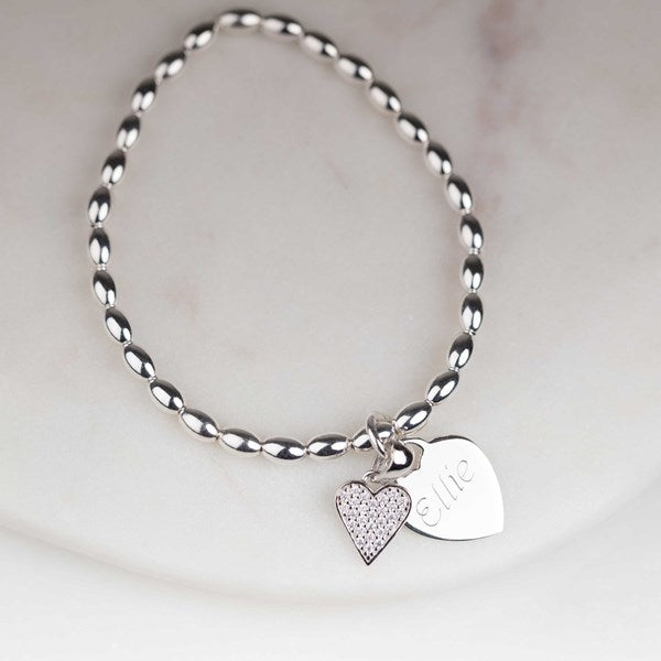 A silver beaded bracelet with two heart-shaped charms, one textured and one engraved with the name 'Ellie', displayed on a light marble surface.