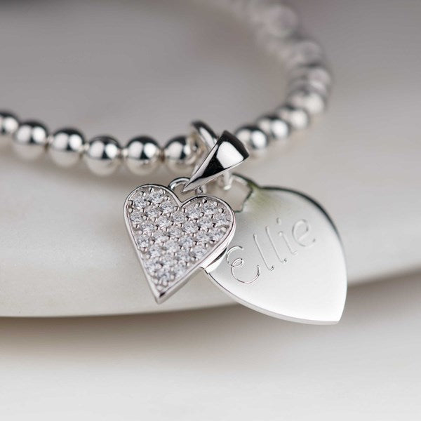 A silver beaded bracelet with two heart-shaped charms, one pavé-set with clear stones and the other engraved with the name 'Ellie', displayed on a light cream-colored surface.