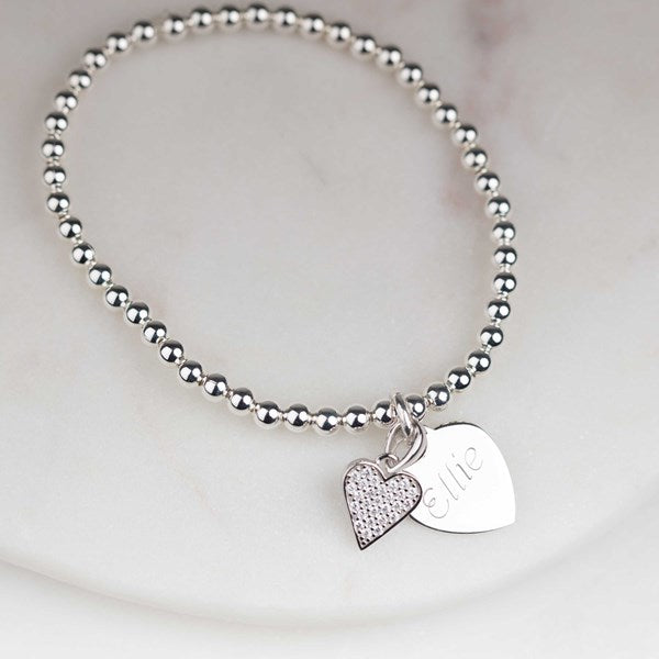 A silver beaded bracelet with two heart-shaped charms, one with cz stones and one engraved with the name 'Elie', displayed on a light marble surface.