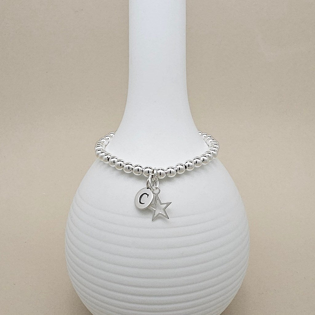 A silver beaded bracelet with a letter 'C' initial pendant and a star charm, displayed on a white ribbed ceramic vase against a beige background.