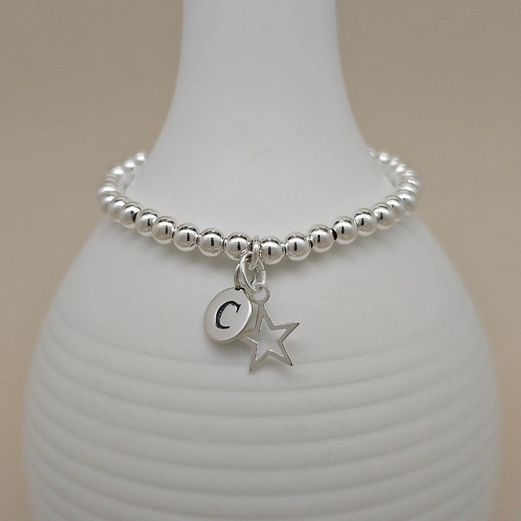 A silver beaded bracelet with a dangling initial 'C' charm and a star charm, displayed on a white ribbed vase against a warm beige background.