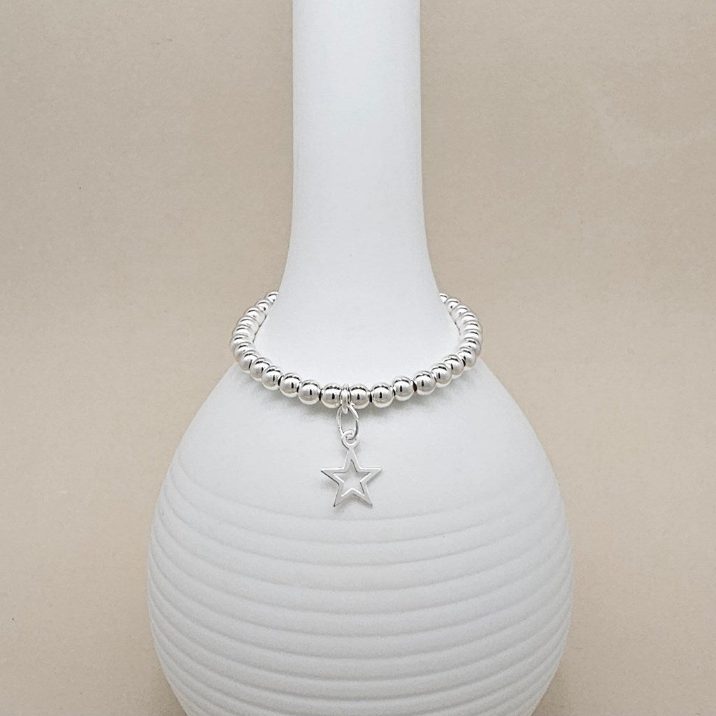 A silver beaded bracelet with a dangling star charm, displayed on a white ribbed ceramic vase against a beige background.