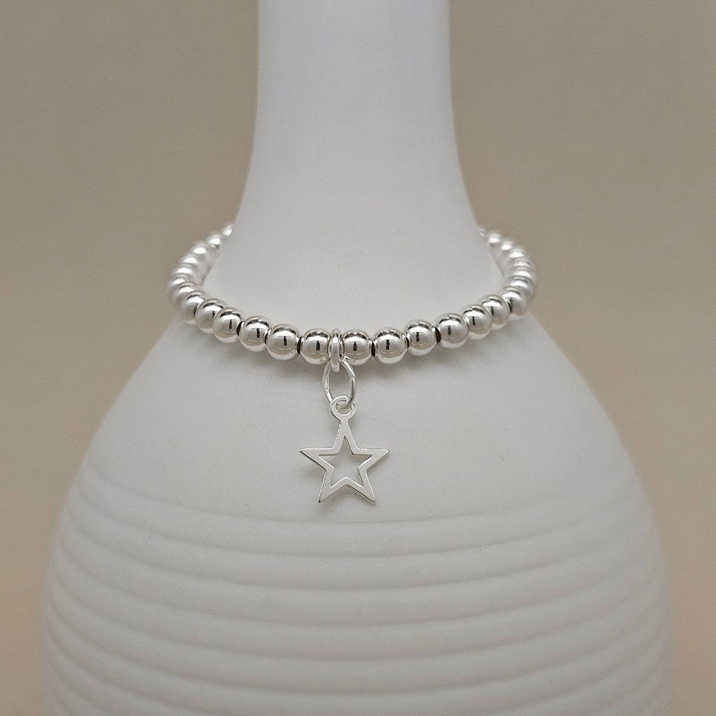 A silver beaded bracelet with a dangling open star charm, displayed on a white ribbed ceramic vase against a soft beige background.