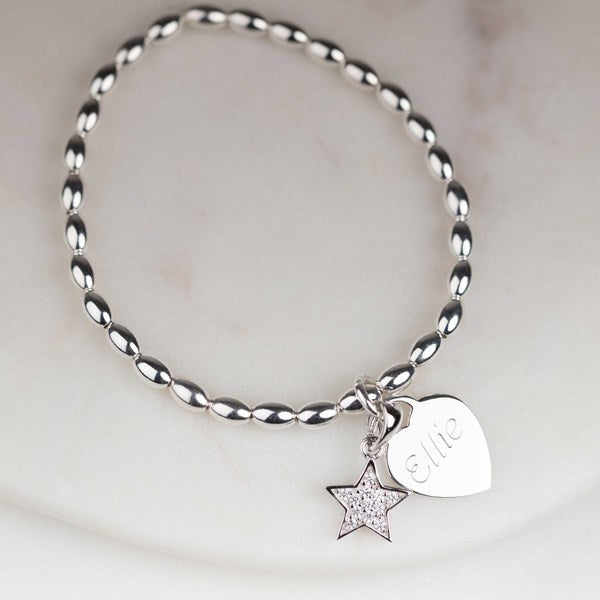 A silver beaded bracelet with two charms, a star charm encrusted with small clear stones and a heart charm engraved with the name 'Ellie', displayed on a light marble surface.