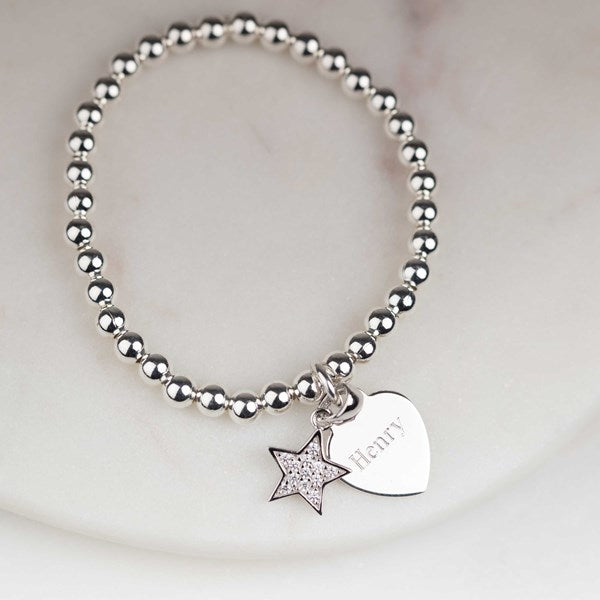 A silver beaded bracelet with two charms: a star charm encrusted with small clear stones and a heart charm engraved with the name 'Henry', displayed on a light marble surface.