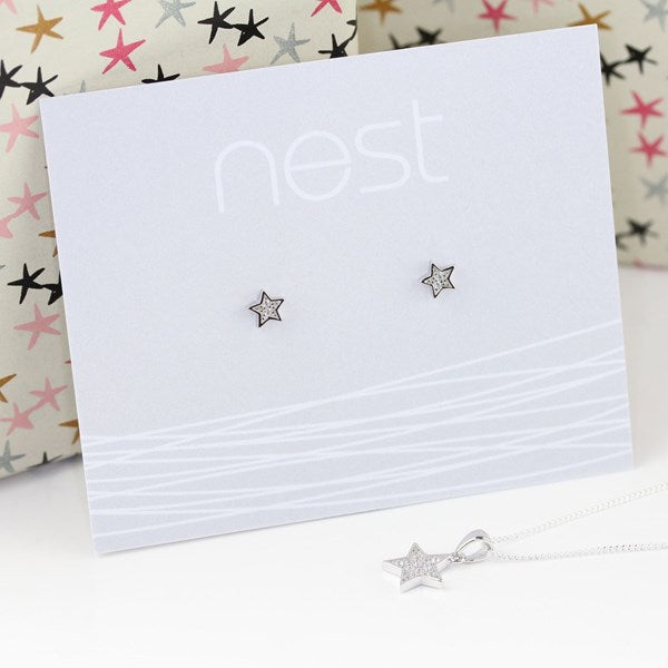 Sterling silver star stud earrings on a white display card with the brand name 'nest', with a matching silver star necklace in the foreground and a patterned gift box in the background.