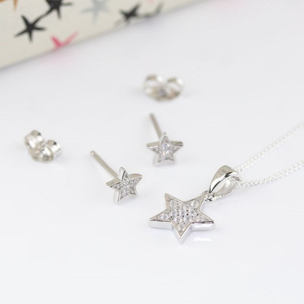 Silver star-shaped jewelry set featuring a pendant necklace, stud earrings, and a small star ring, displayed on a clean white surface with a blurred patterned box in the background.
