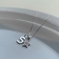 A silver necklace with a box chain, featuring a lowercase letter 's' pendant and a small star charm studded with clear rhinestones, resting on a light gray ceramic dish.