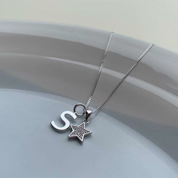 A silver necklace with a box chain, featuring a lowercase letter 's' pendant and a small star charm studded with clear rhinestones, resting on a light gray ceramic dish.
