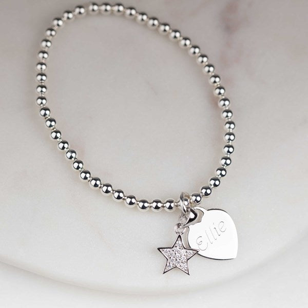 A silver beaded bracelet with a dangling star charm and a heart-shaped tag engraved with the name 'Elite', displayed on a light marble surface.