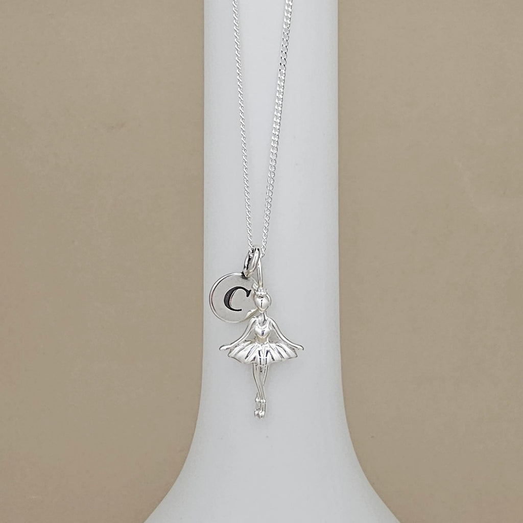 A silver ballerina pendant necklace with a letter charm, displayed on a white jewelry bust against a beige background.