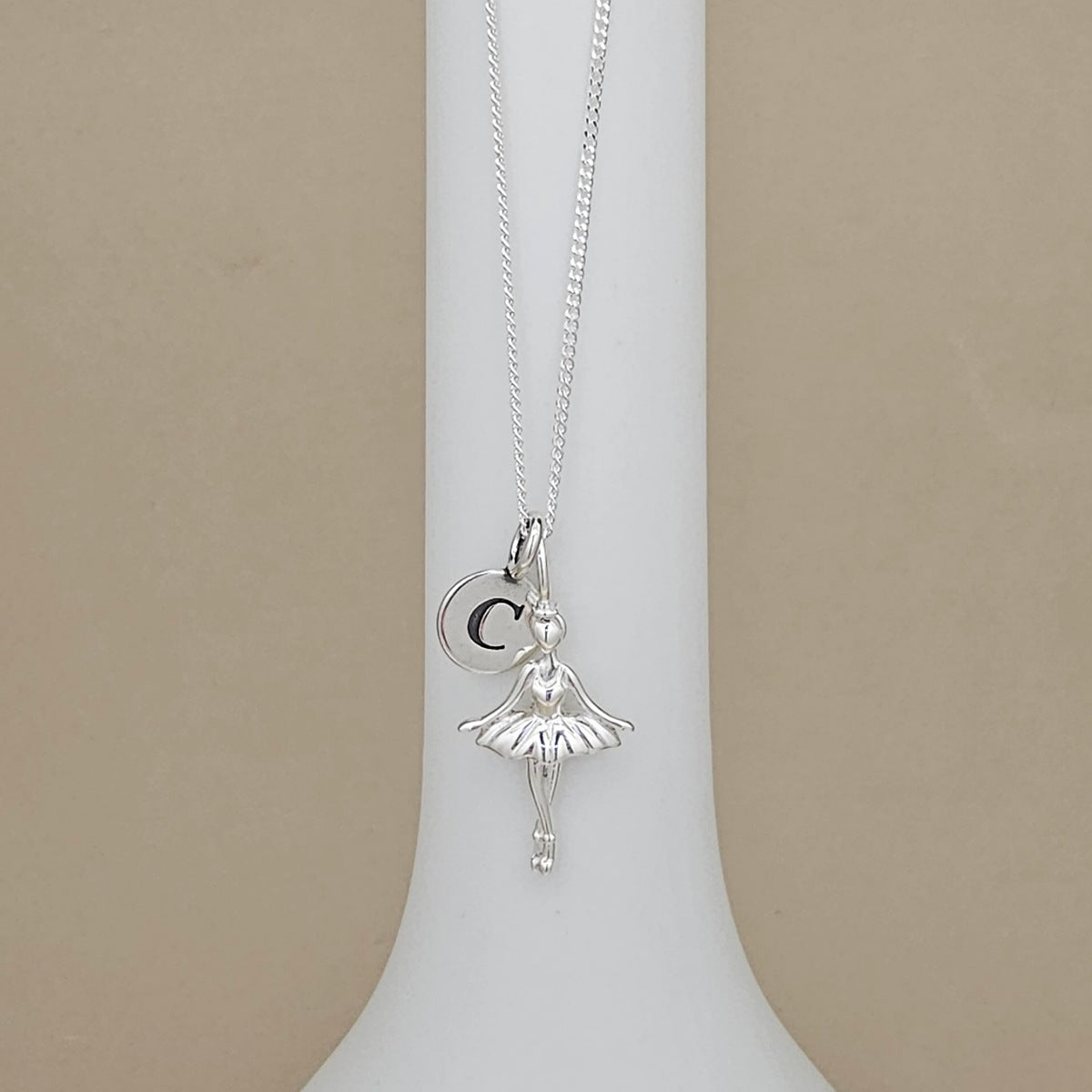 A silver ballerina pendant necklace with a letter charm, displayed on a white jewelry bust against a beige background.