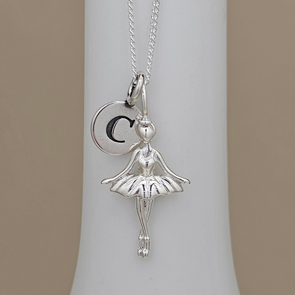 A silver ballerina pendant necklace with a matching initial 'C' charm, displayed on a white cylindrical stand against a neutral beige background.