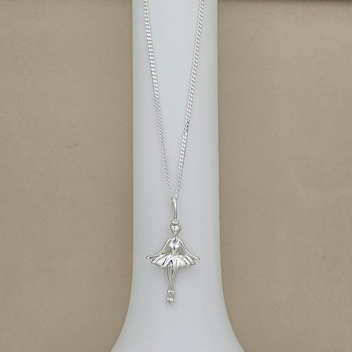 A silver ballerina pendant necklace displayed on a white cylindrical stand against a neutral beige background.