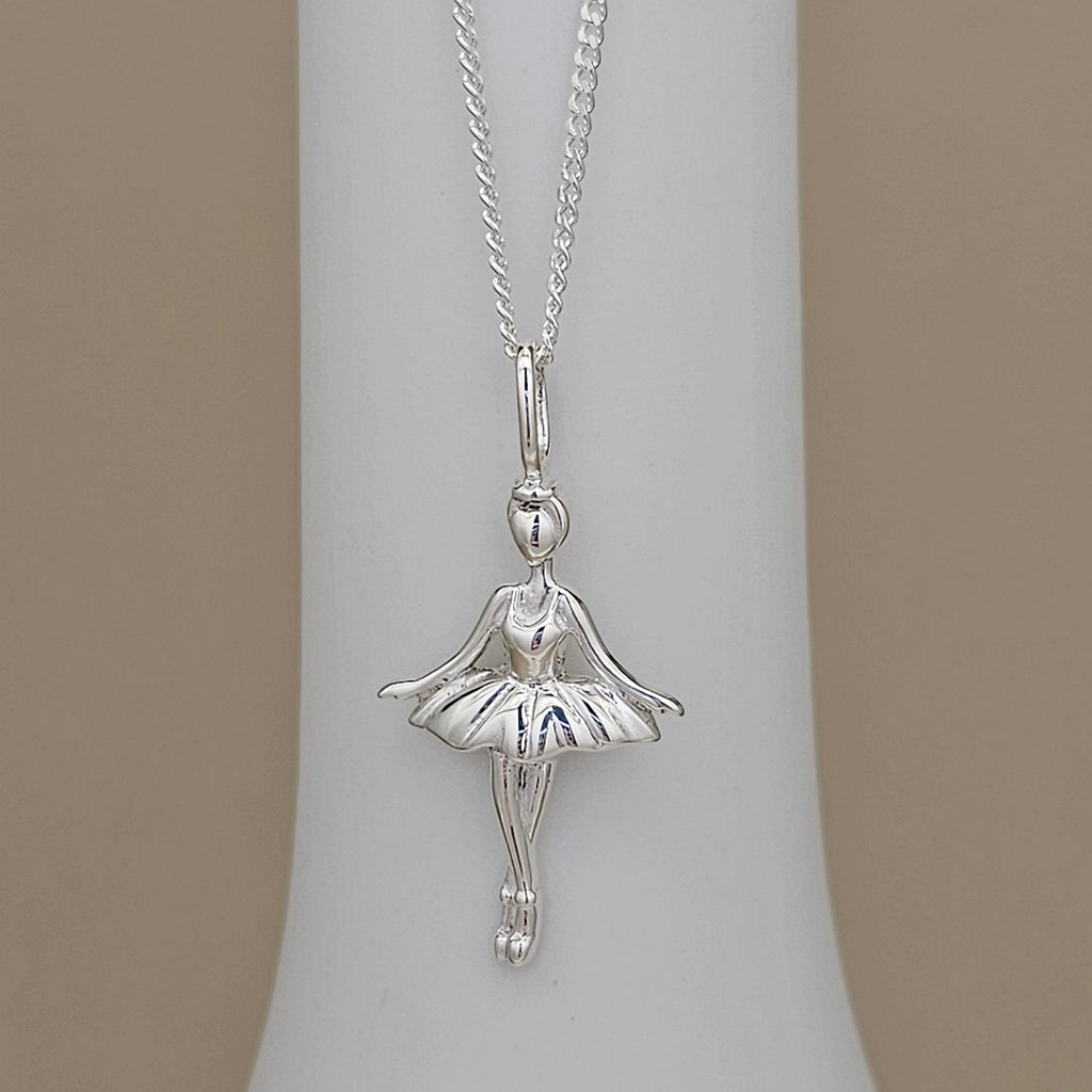 A silver ballerina pendant necklace displayed on a white cylindrical jewelry stand against a warm beige background.