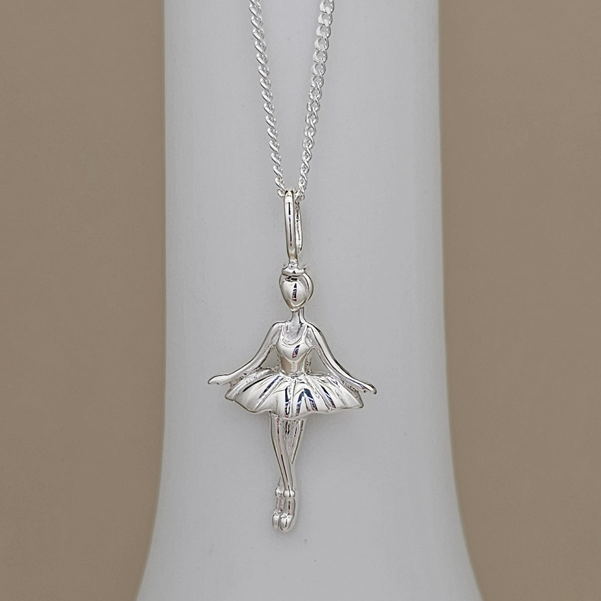 A silver ballerina pendant necklace displayed on a white cylindrical jewelry stand against a warm beige background.