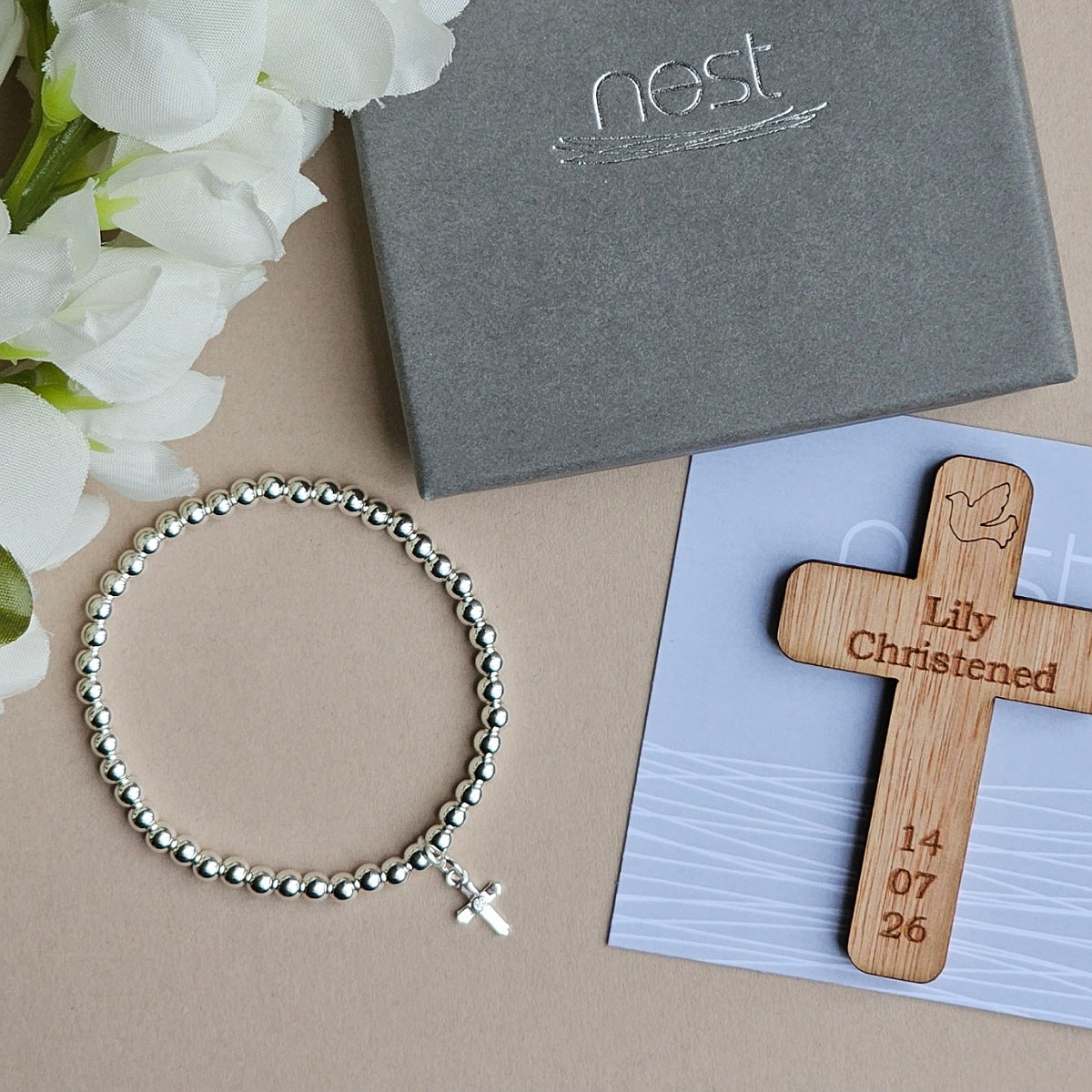 Bracelet, wooden cross with engraving, and 'nest' box on a beige surface with flowers.