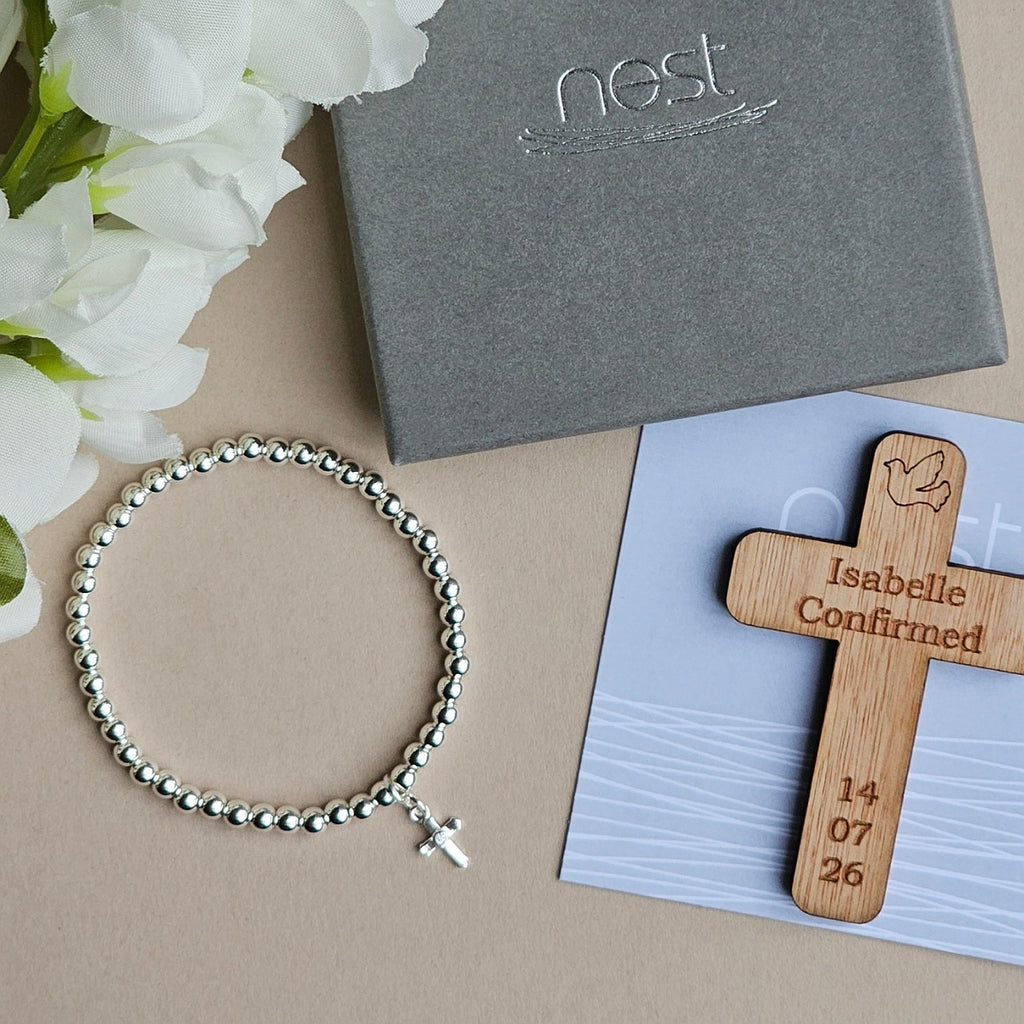 A silver beaded bracelet with a small cross charm is arranged on a beige surface beside white flowers, a gray Nest gift box, and a wooden confirmation cross engraved with the name Isabelle and a date, placed on a light blue card.
