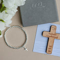A silver beaded bracelet with a small cross charm is arranged on a beige surface beside white flowers, a gray Nest gift box, and a wooden confirmation cross engraved with the name Isabelle and a date, placed on a light blue card.