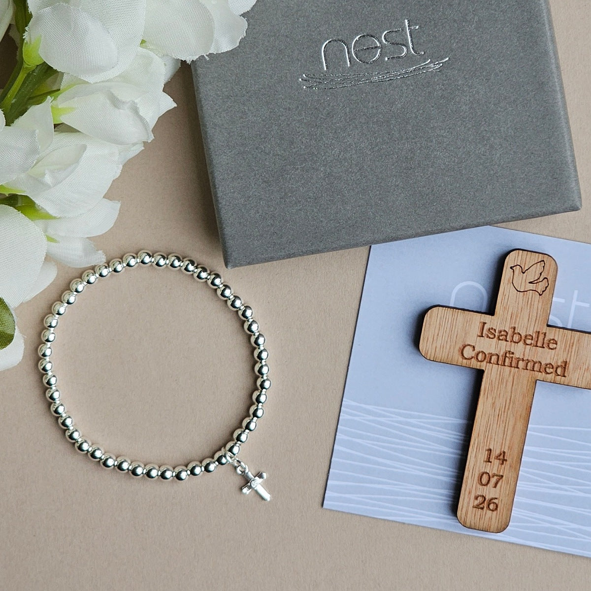 A silver beaded bracelet with a small cross charm is arranged on a beige surface beside white flowers, a gray Nest gift box, and a wooden confirmation cross engraved with the name Isabelle and a date, placed on a light blue card.
