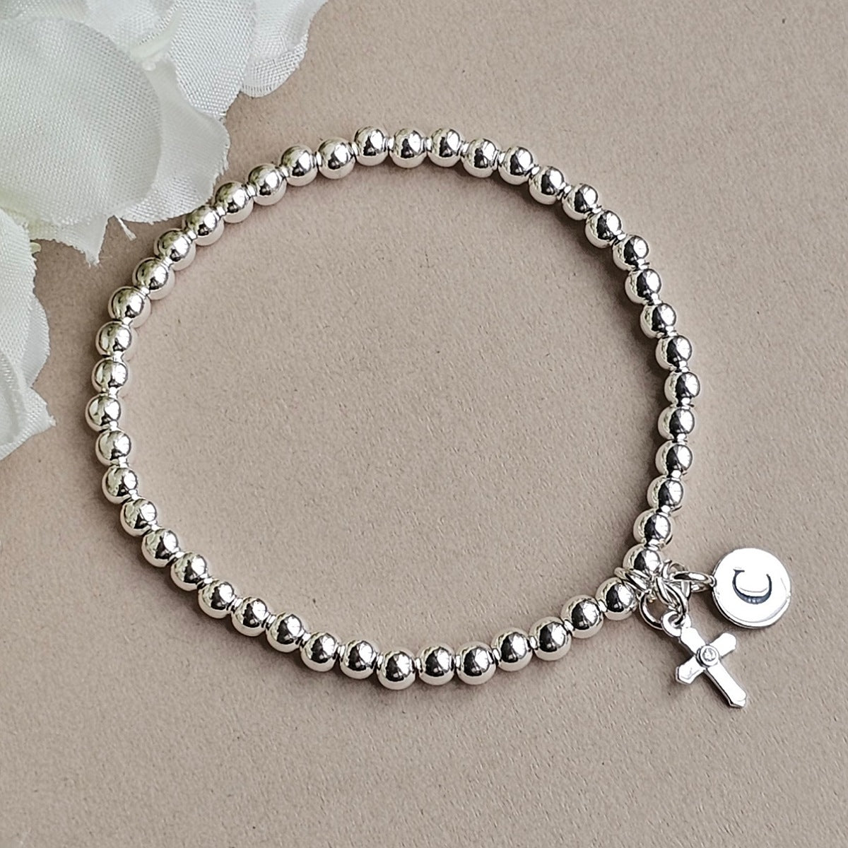 A silver beaded bracelet with a small charm featuring a cross and a round disc engraved with a crescent moon, displayed on a neutral beige background with a white flower in the corner.